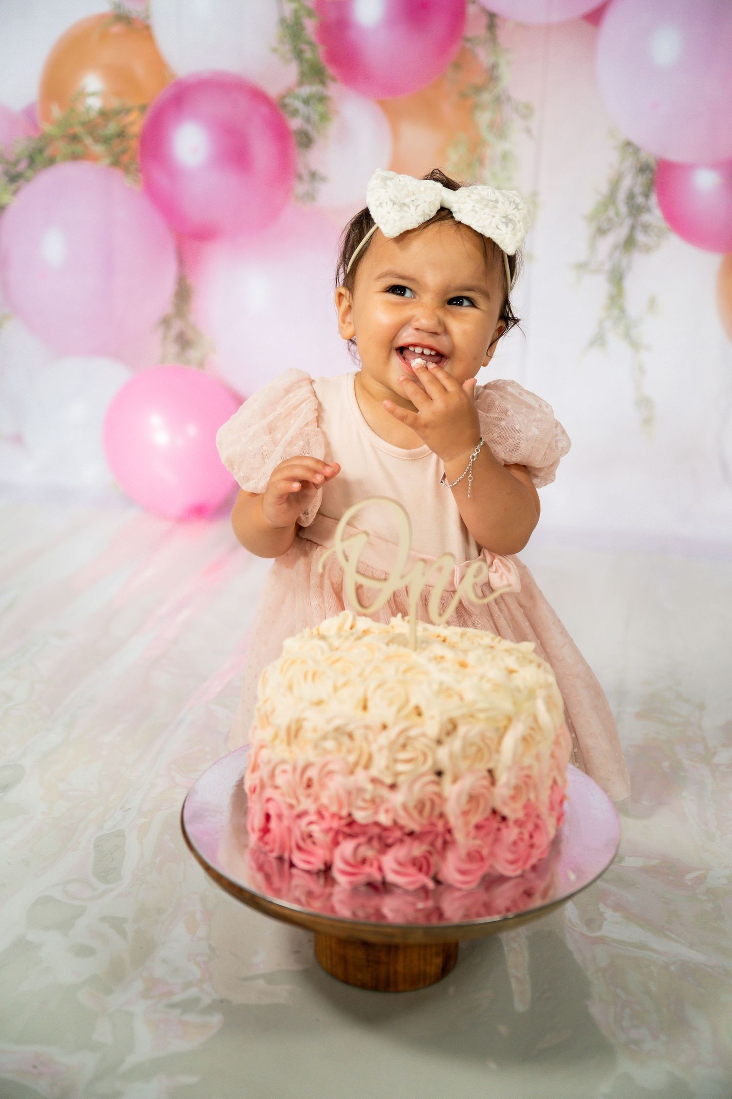 First birthday cake smash photo of a smiling baby girl with pink balloons – colourful and fun milestone photography in Tweed Heads.