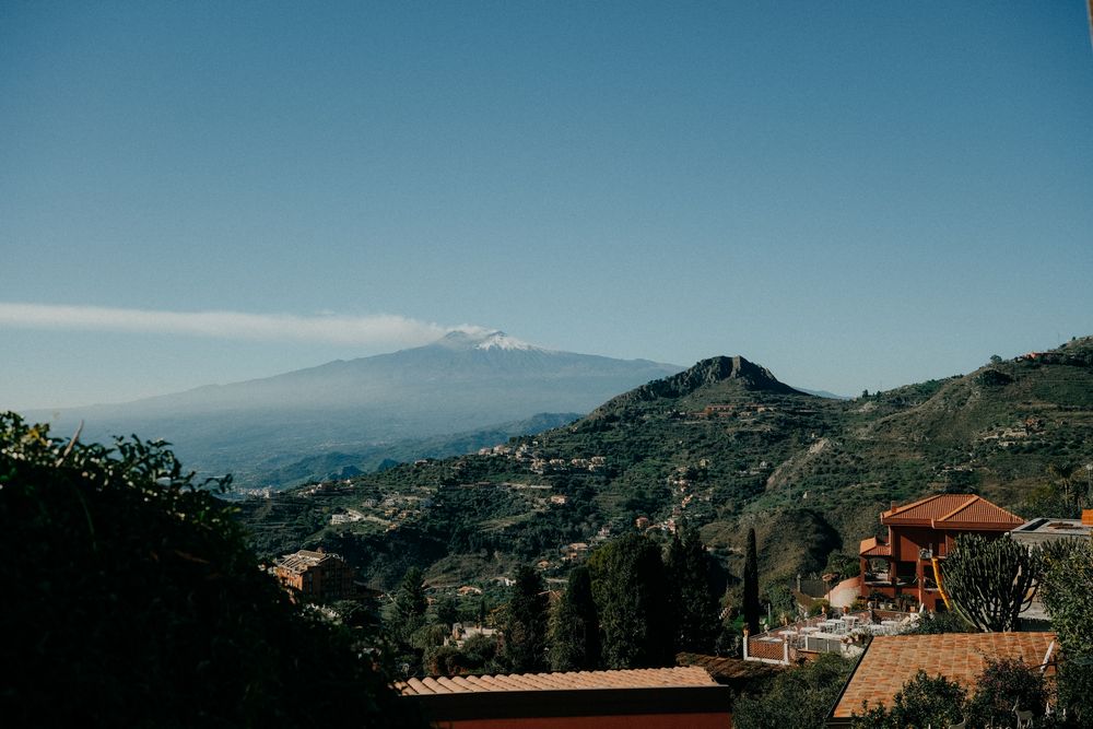 Panoramic view of a Mediterranean coastal town with terracotta roofs nestled in mountainous terrain under blue skies.