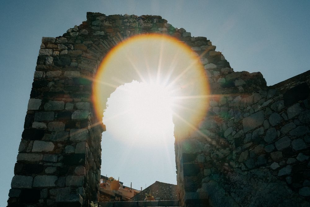 Bright sunlight bursts through a stone archway in ancient ruins creating a dramatic lens flare effect.