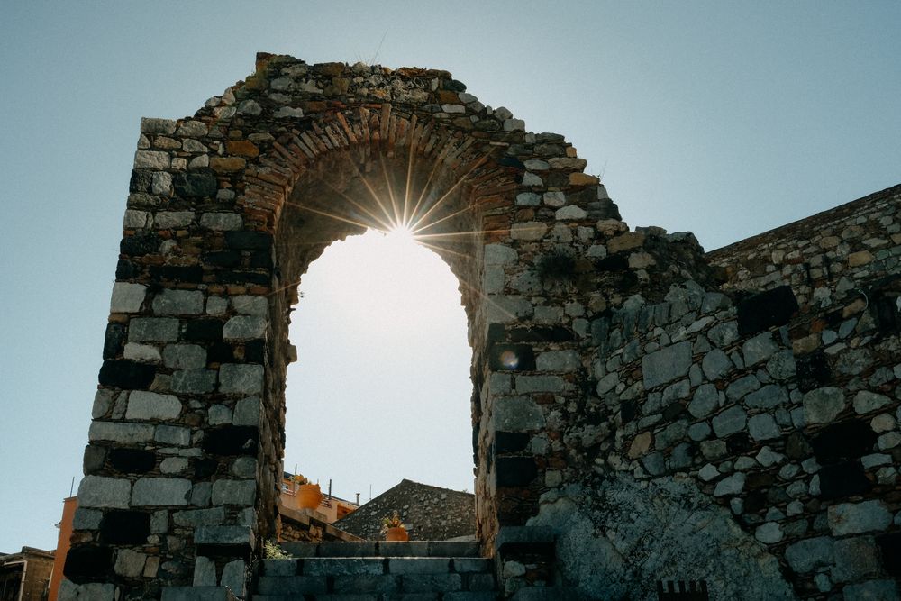Stone archway ruins silhouetted against a bright sunburst in a clear blue sky.