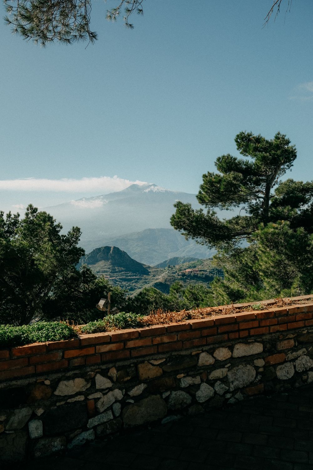 Stone wall overlooks a scenic mountain landscape with pine trees and blue skies in the Mediterranean.