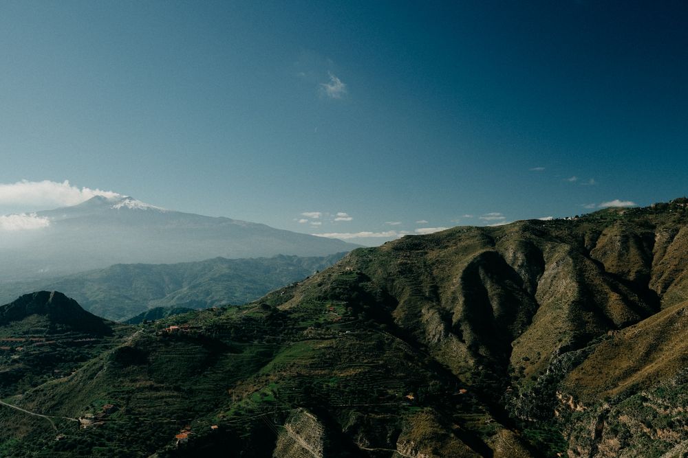 Dramatic mountain landscape with terraced hillsides and deep valleys under a bright blue sky with scattered clouds.