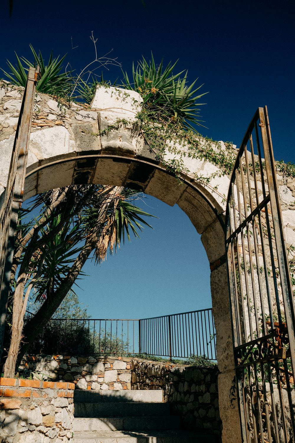 Stone archways with wrought iron railings line a Mediterranean garden path under palm trees and bright blue sky.