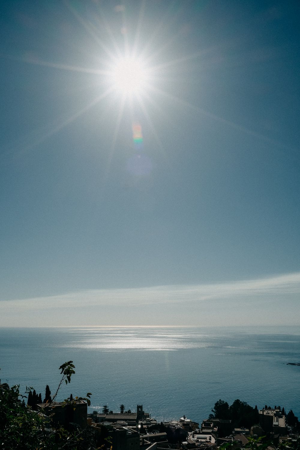 Bright sun shines over calm ocean waters with coastal buildings and palm trees silhouetted against the horizon.