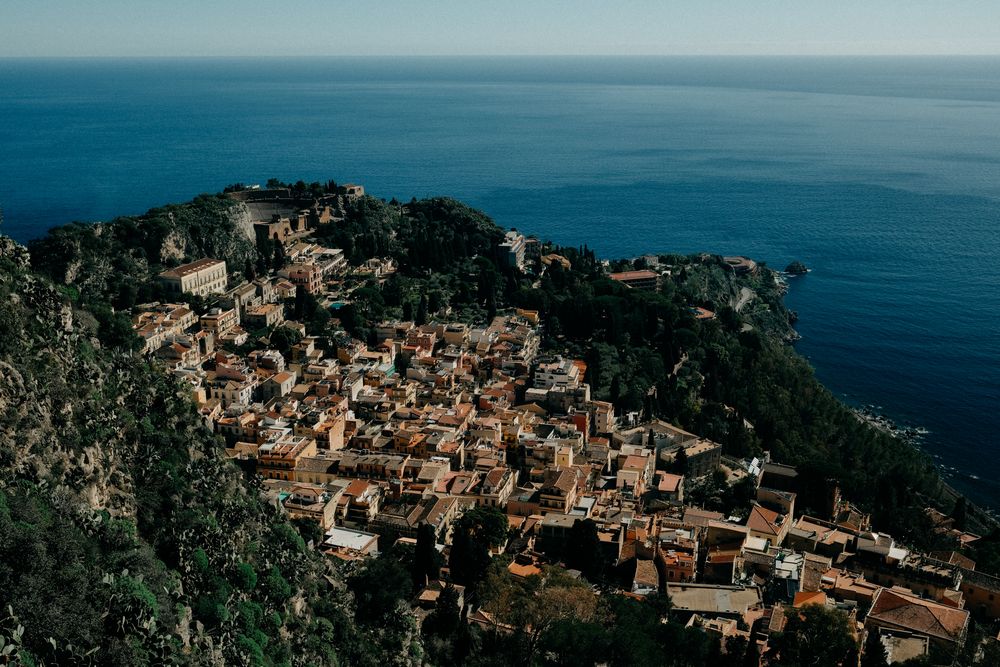 Panoramic aerial view of a coastal Mediterranean town with orange terracotta roofs nestled along cliffside overlooking blue sea.