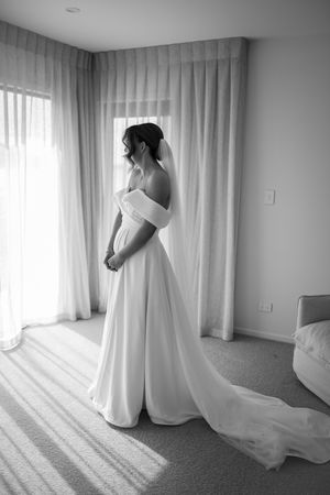 Sunlit bridal preparation room with flowing white wedding dress and sheer curtains creating soft shadows.