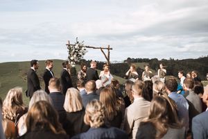 An outdoor wedding ceremony takes place under a rustic wooden arch on a hillside with guests gathered to witness the occasion.