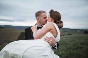 Romantic wedding couple embracing on hilltop against moody sky with bride's veil flowing in wind.