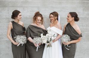 Bridal party in olive green dresses and white wedding gown pose together holding baby's breath bouquets against grey wall.