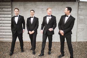 Groomsmen in black tuxedos sitting on a wooden bench sharing laughs during wedding photos.