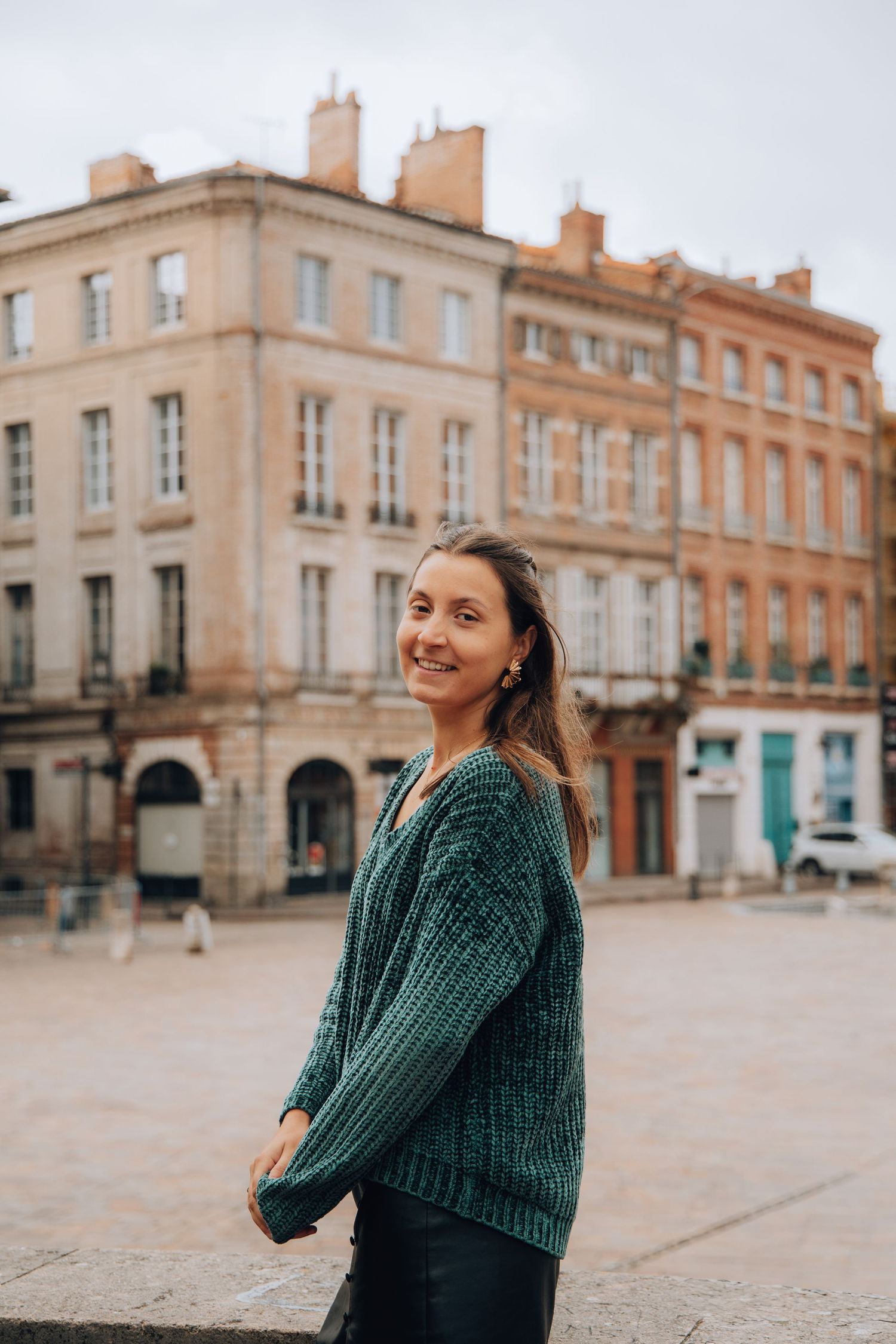Smiling woman in green knit sweater standing in European city square with classic buildings.