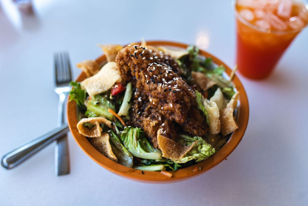 A crispy sesame-crusted chicken salad served in an orange bowl with mixed greens and a bright cocktail on the side.