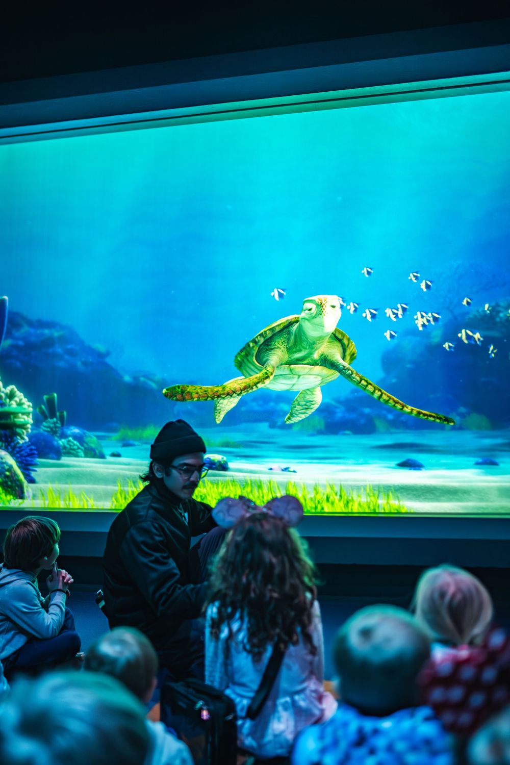 Visitors watch sea turtles swim by in a large aquarium display with vibrant blue lighting and floating bubbles. Crush's Turtle Talk