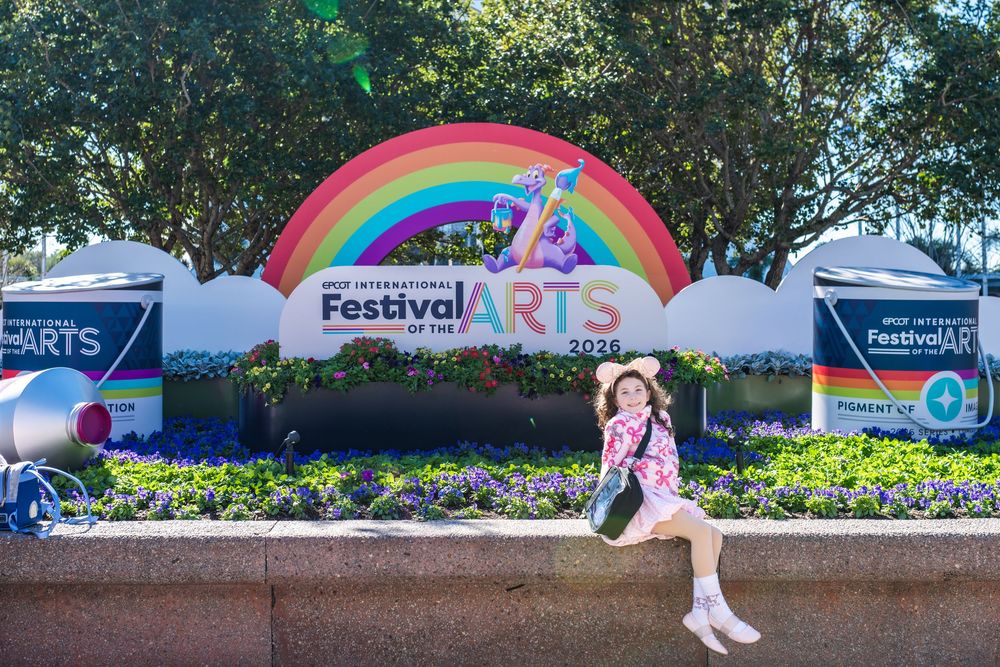 A rainbow art installation stretches across a pathway with festival banners and trees in the background. Epcot Festival Of The Arts