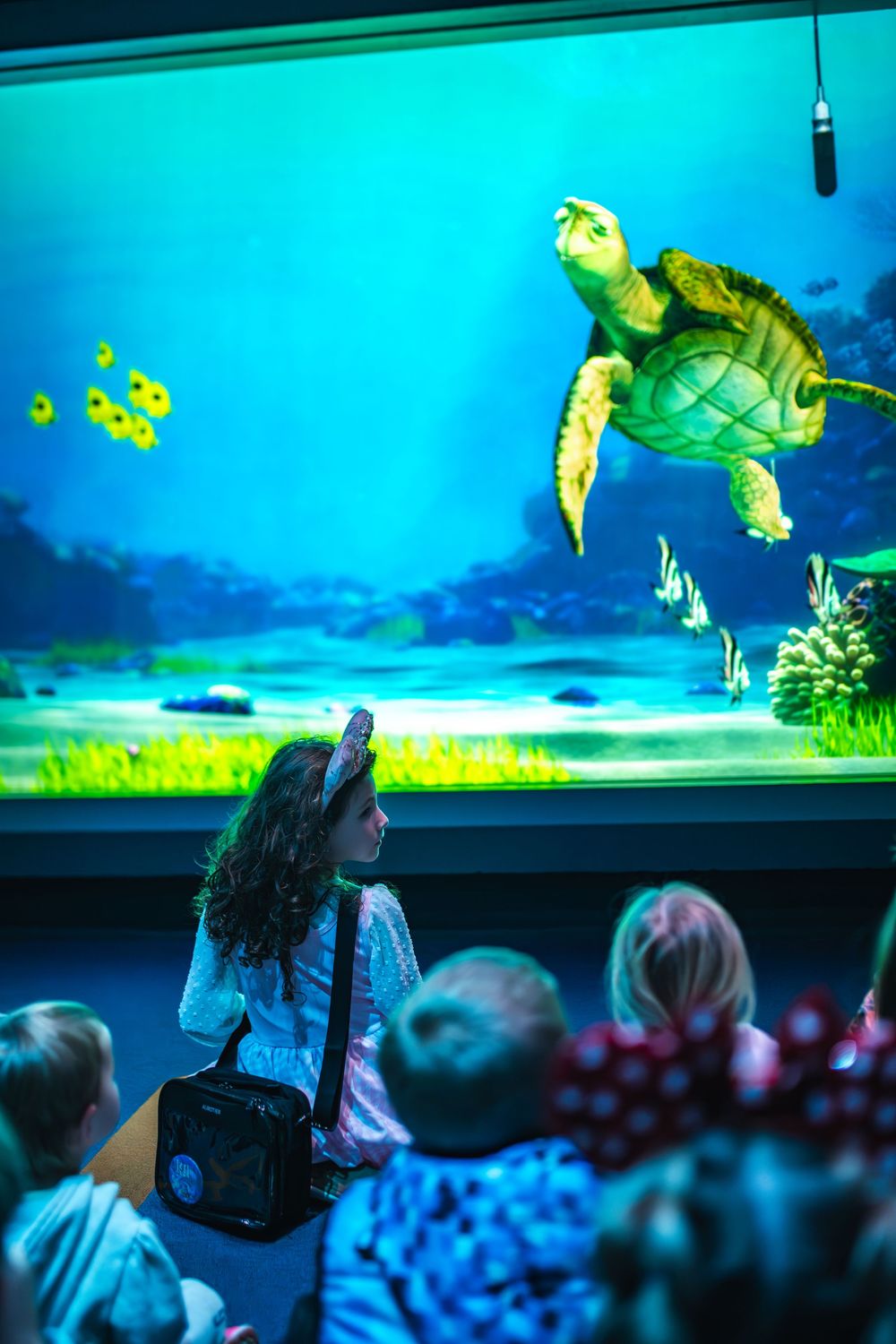 Visitors watch vibrant yellow fish swimming in a large aquarium display with bright turquoise water.