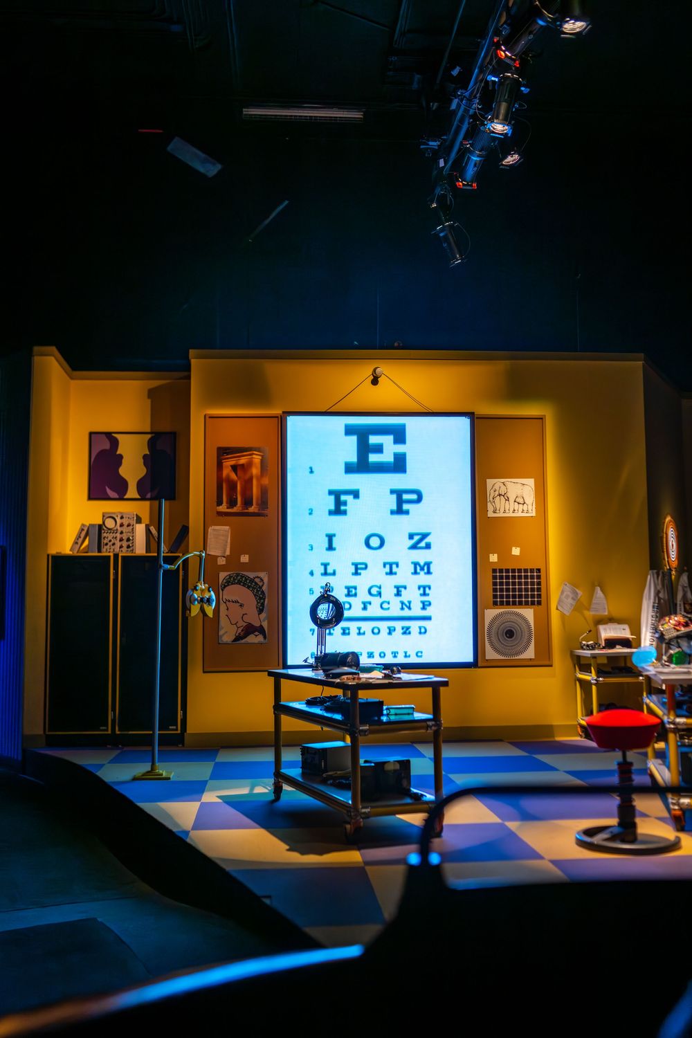 Dimly lit optometry office interior showing eye exam equipment and illuminated eye chart displays on the walls.