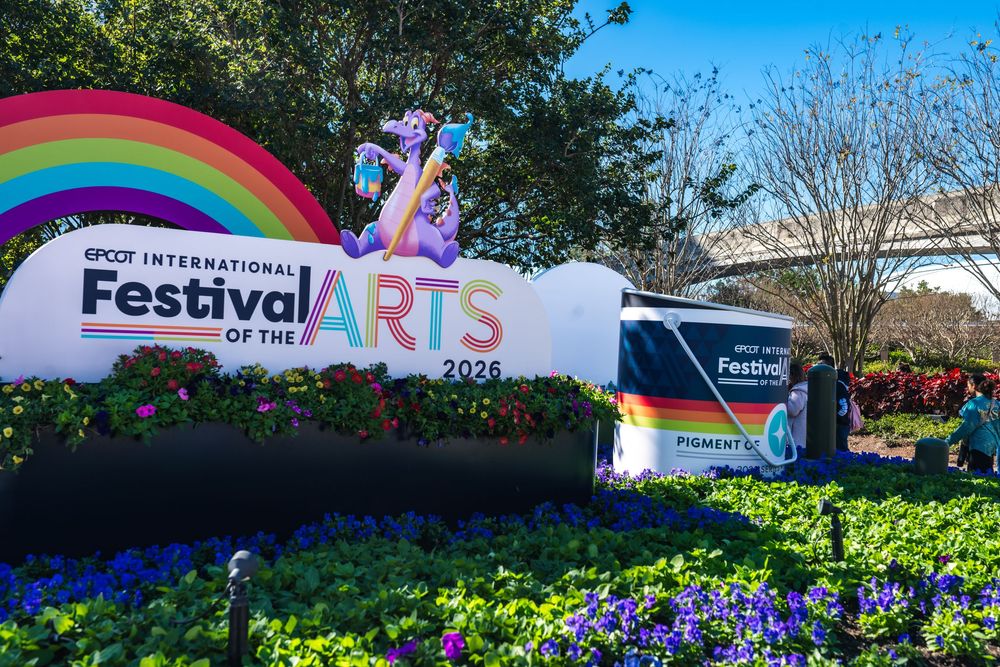 Colorful rainbow banners and festival signage decorate an outdoor arts event under sunny blue skies.