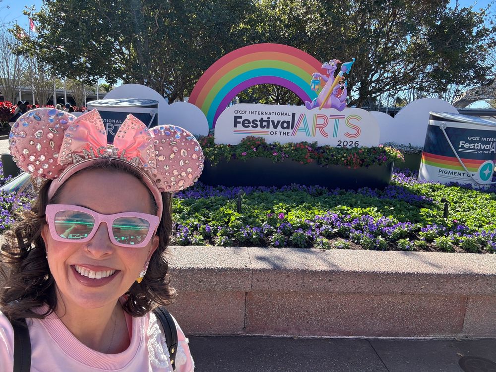 Visitor wearing pink Mickey Mouse ears and sunglasses smiles while enjoying a sunny day at Disney theme park. Epcot Festival Of The Arts