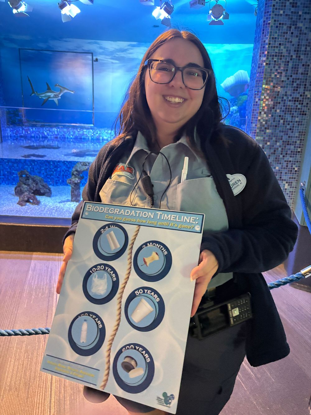 Staff member displays shark embryo timeline educational board in front of blue aquarium tank display.