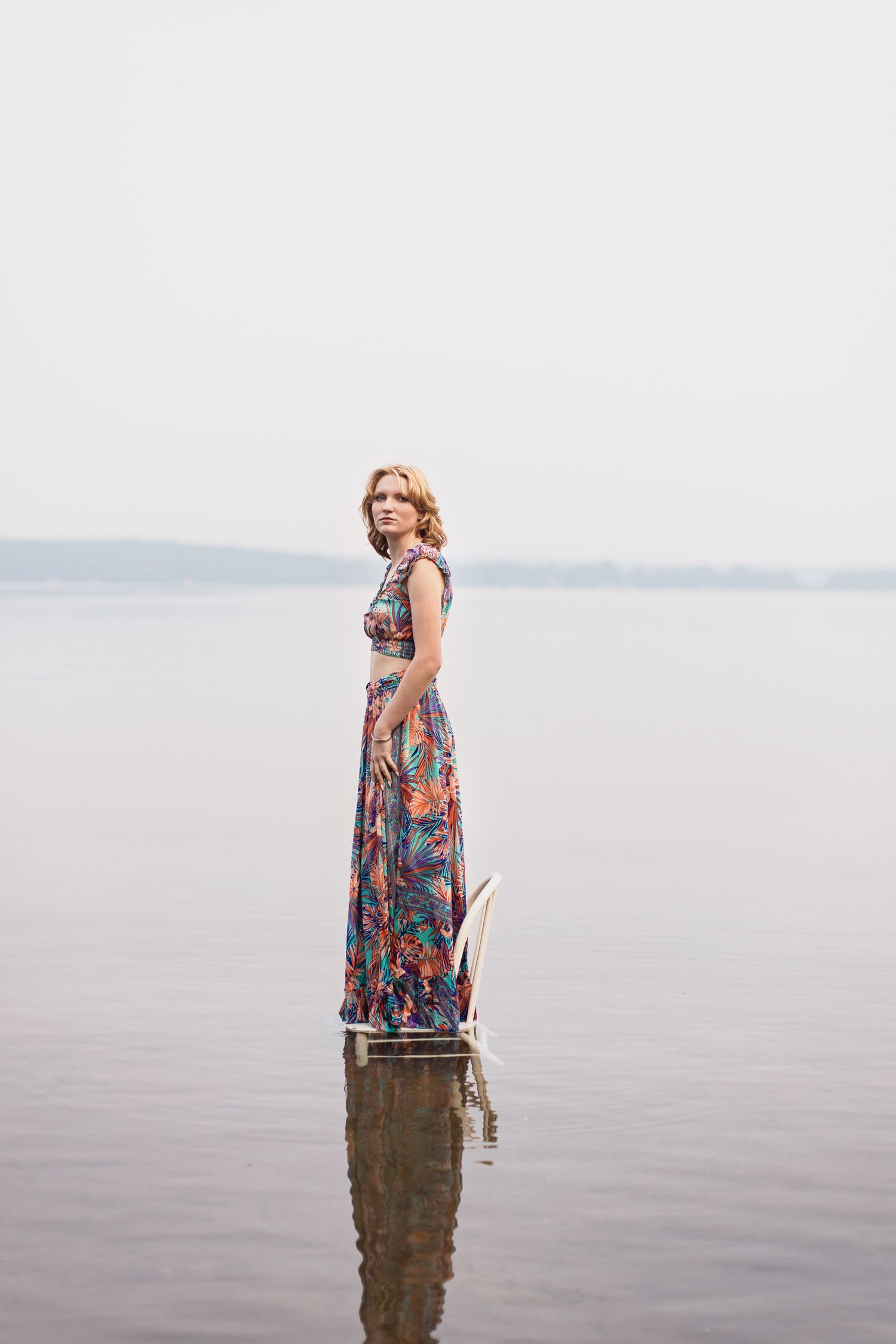 A person in a floral dress stands in shallow water against a misty coastal backdrop creating a serene scene.