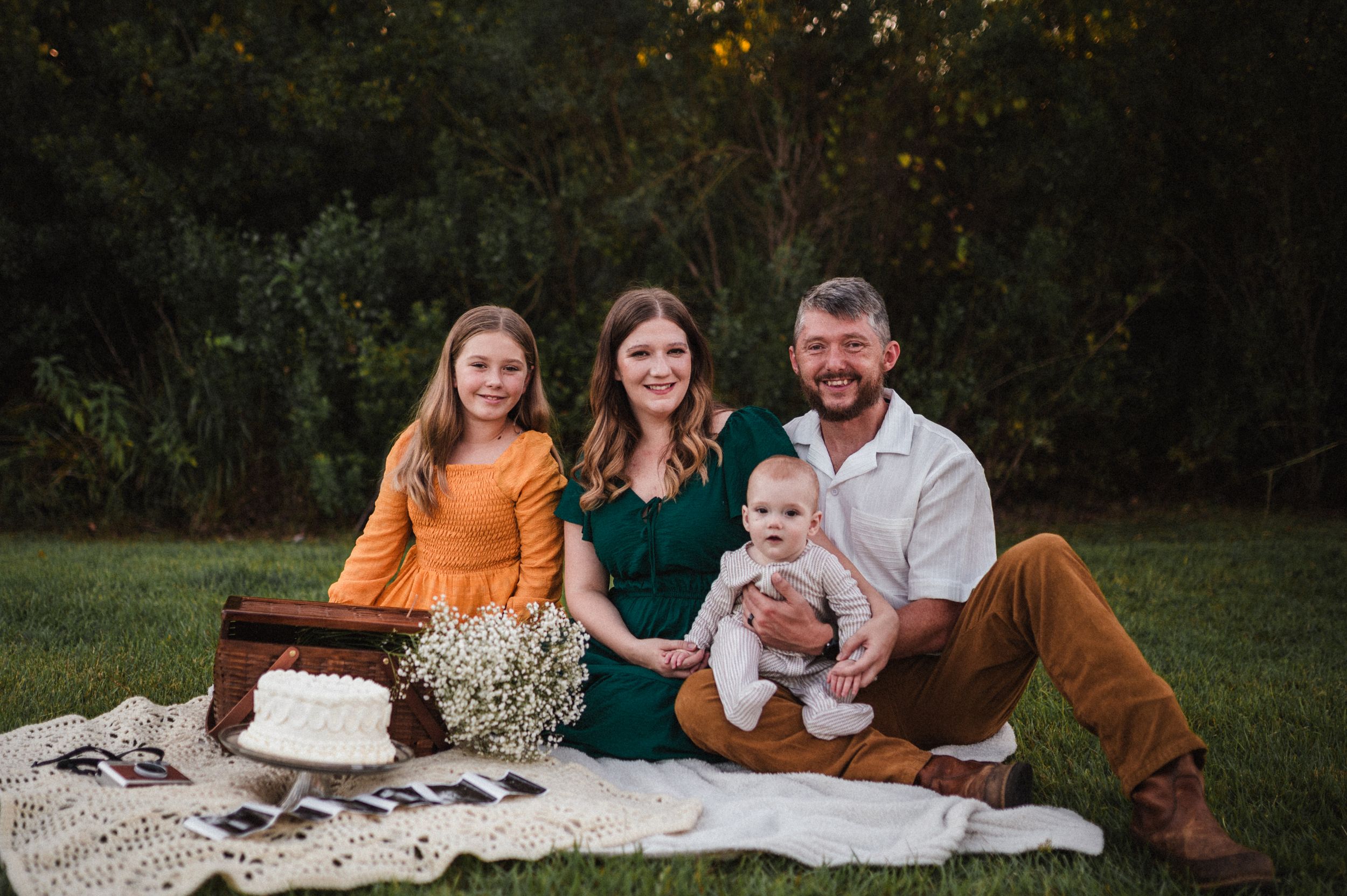 A family sits together on a white blanket during an outdoor photo session in a grassy field at sunset.