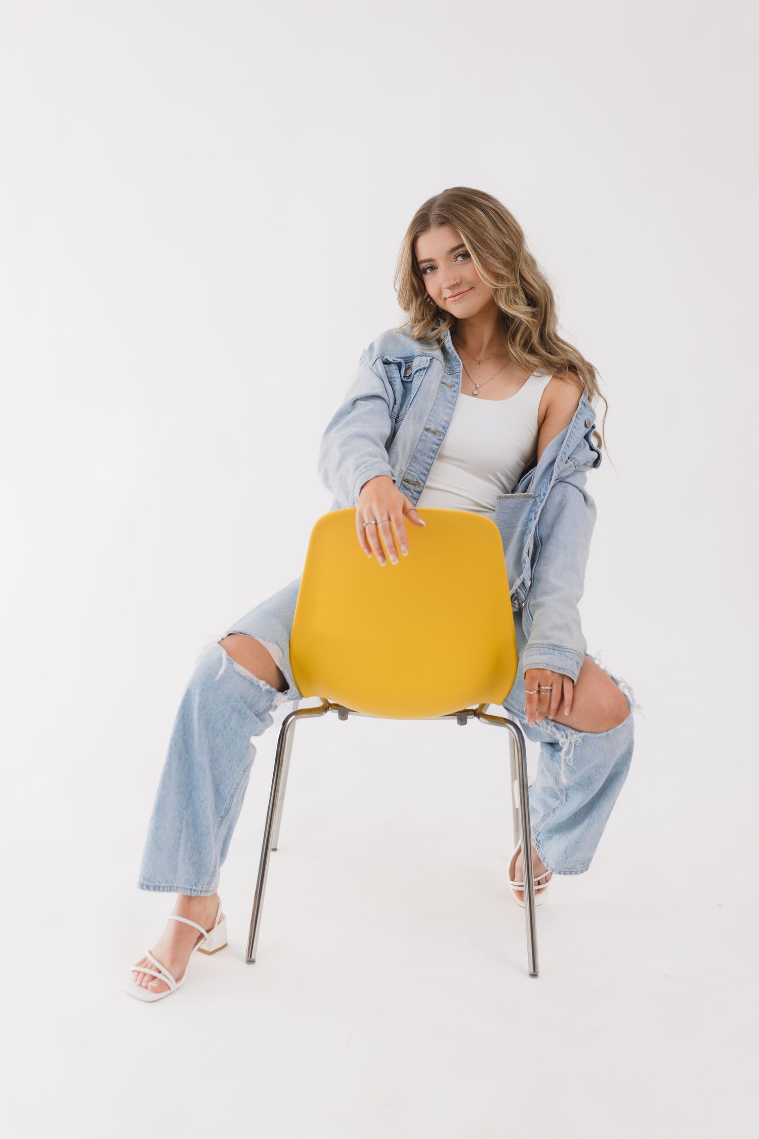 Fashion model in light denim and white top poses creatively on yellow chair against white background.