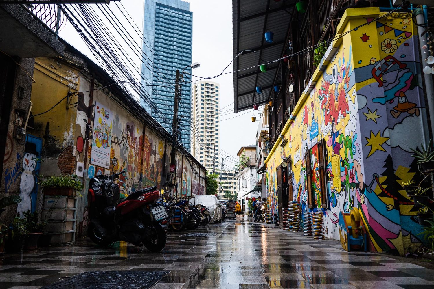 Colorful street art murals line the walls of an urban alleyway with tall buildings in the background on a rainy day.
