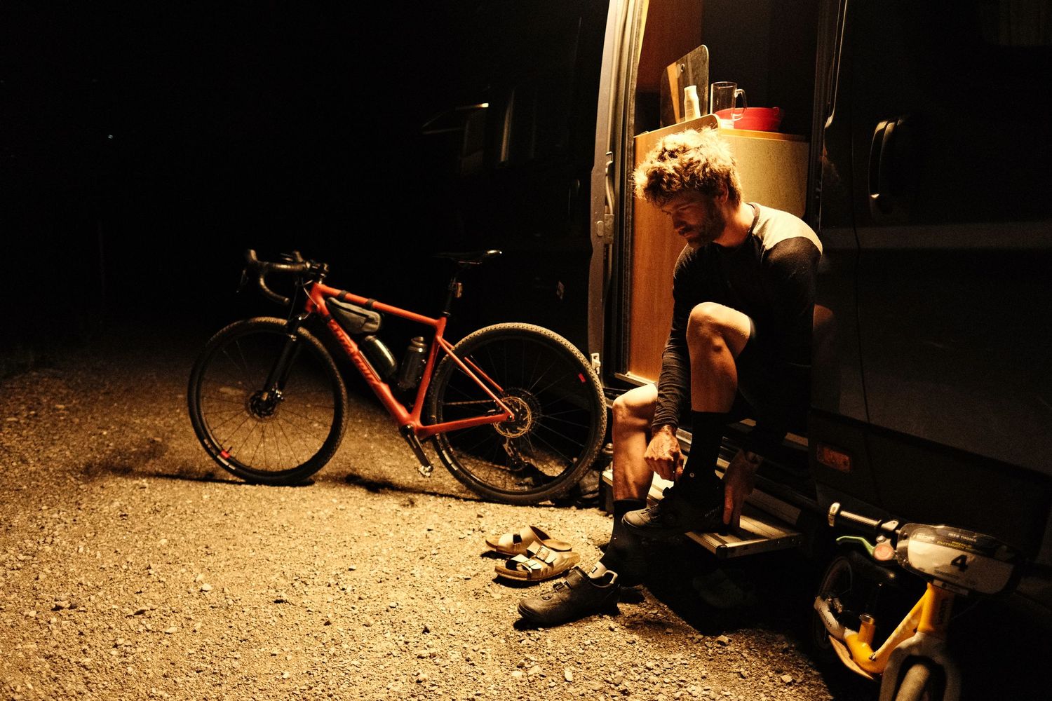 A red bicycle parked outside at night while someone sits in a doorway with equipment under warm lighting.