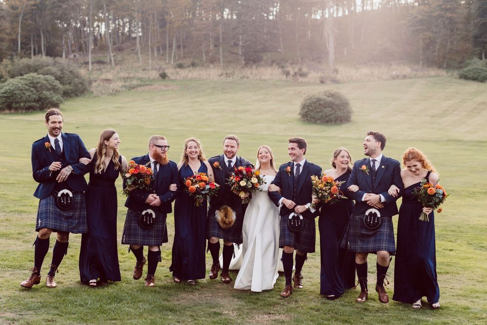 Wedding party in navy blue attire poses playfully on a grassy hillside in autumn.