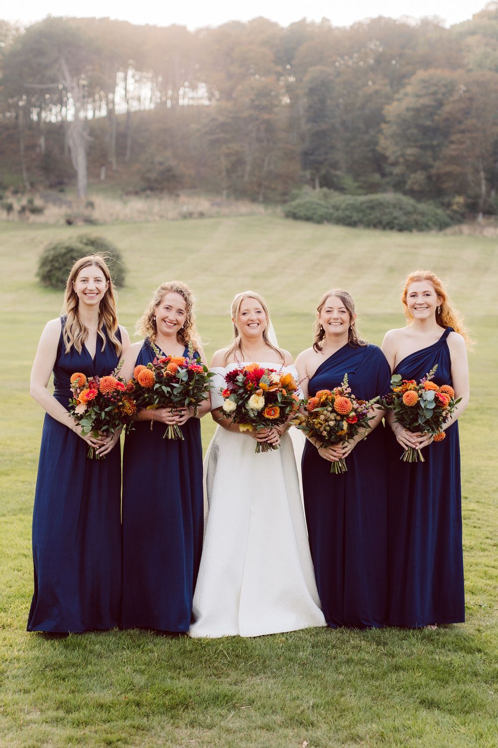 Wedding party in navy blue bridesmaid dresses holding vibrant orange and red fall bouquets on a grassy field.