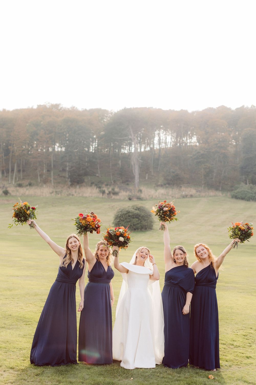 Bridesmaids in navy dresses and bride in white gown pose together in a scenic field with autumn bouquets.