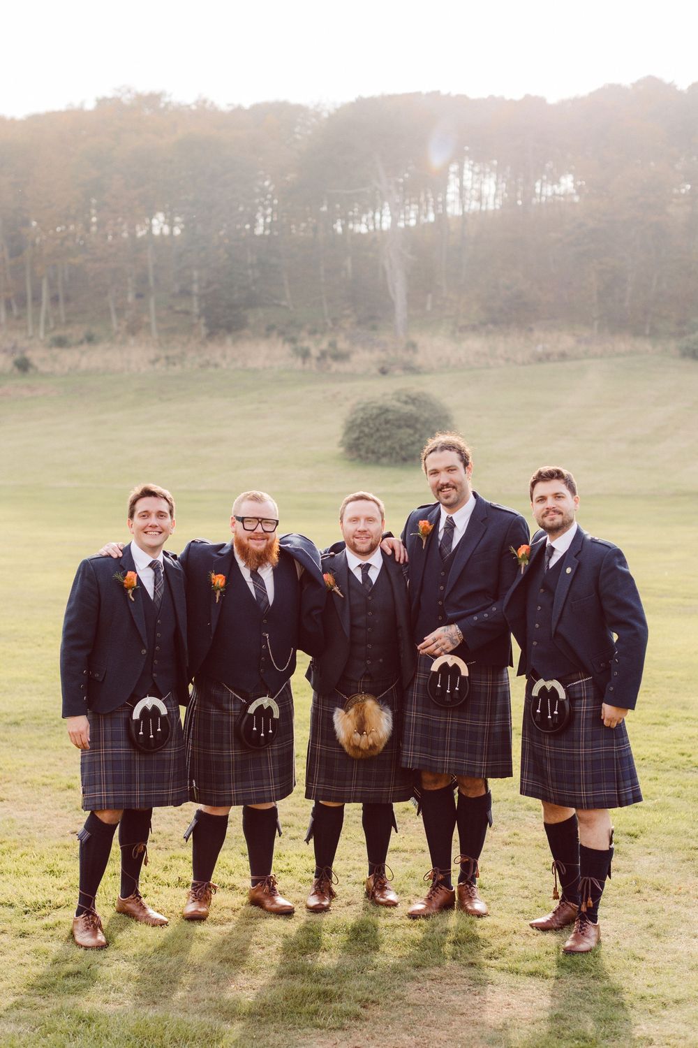 A group of groomsmen in traditional Scottish kilts pose playfully in a field showing their socks.