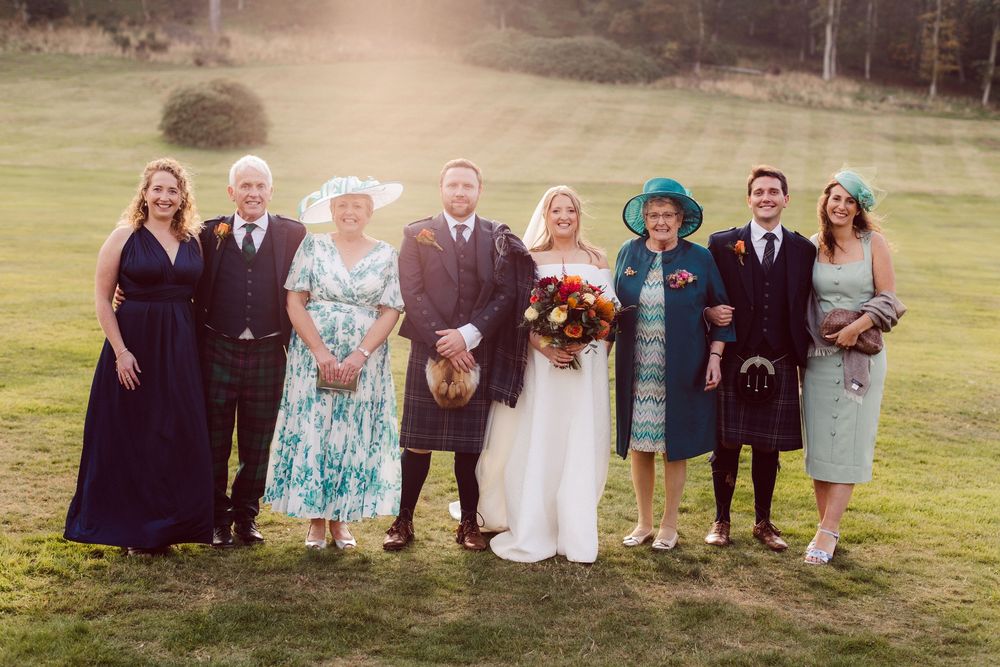 A wedding party group poses together outdoors on a grassy field wearing formal attire in various colors.