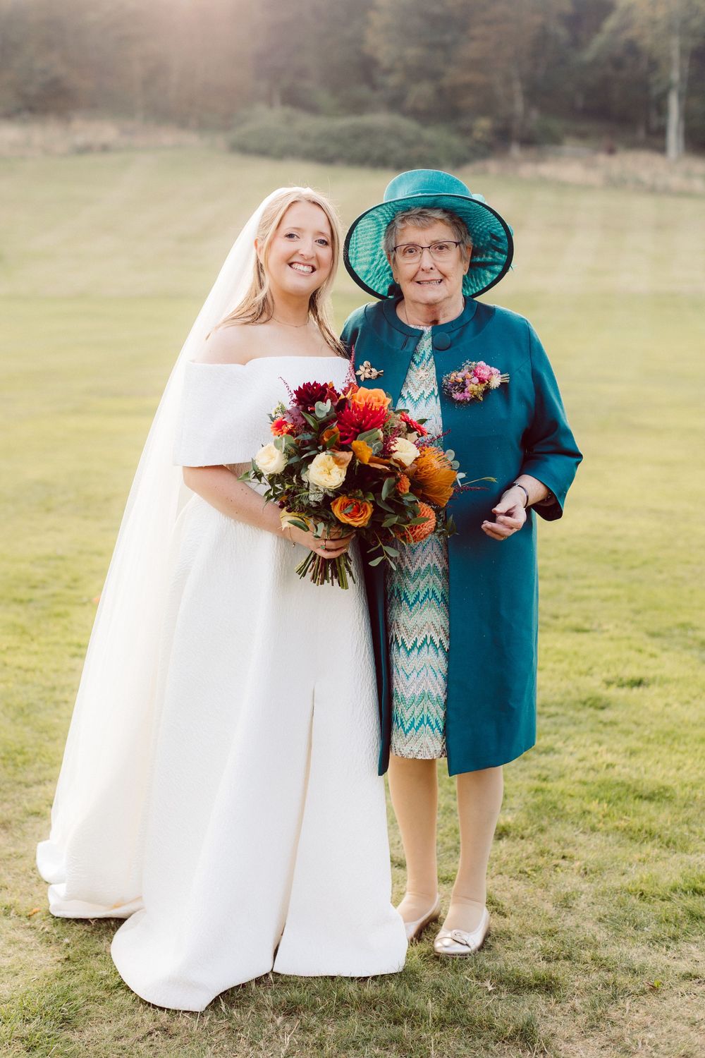 A bride in a white off-shoulder gown stands with an older woman in a teal dress and hat holding colorful fall bouquets.
