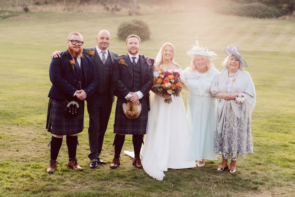A group of wedding guests pose together on a grassy field during an outdoor celebration.