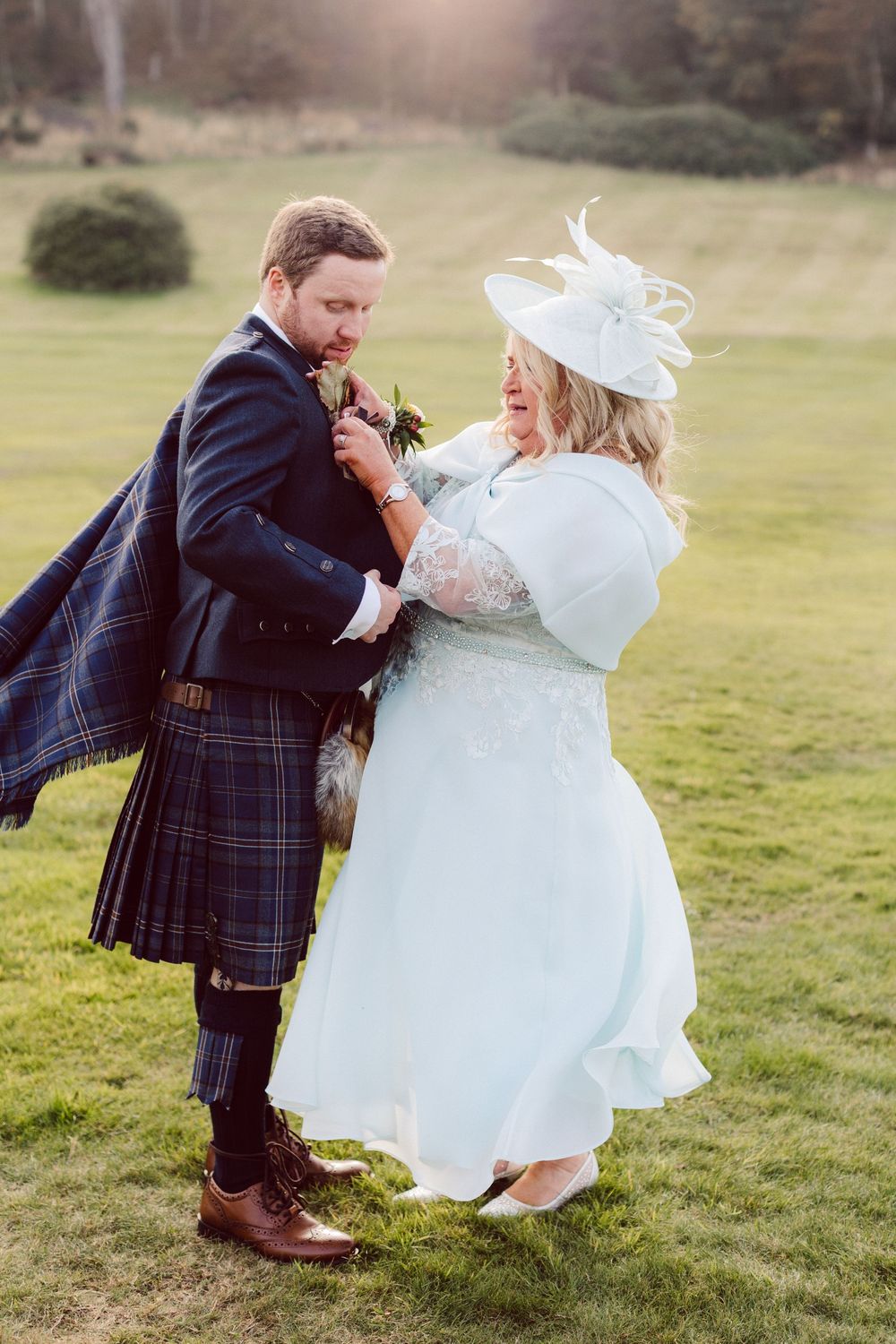 A couple in wedding attire dances playfully in a grassy field, with the groom wearing a traditional Scottish kilt.