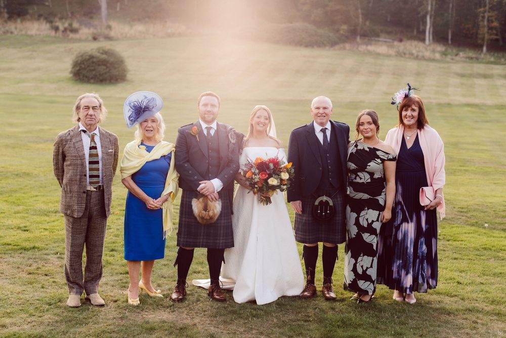 Wedding group photo with guests arranged in a line outdoors on a grassy hillside.