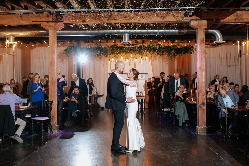 Wedding guests watch as newlyweds dance in a rustic venue with exposed beams and string lights.