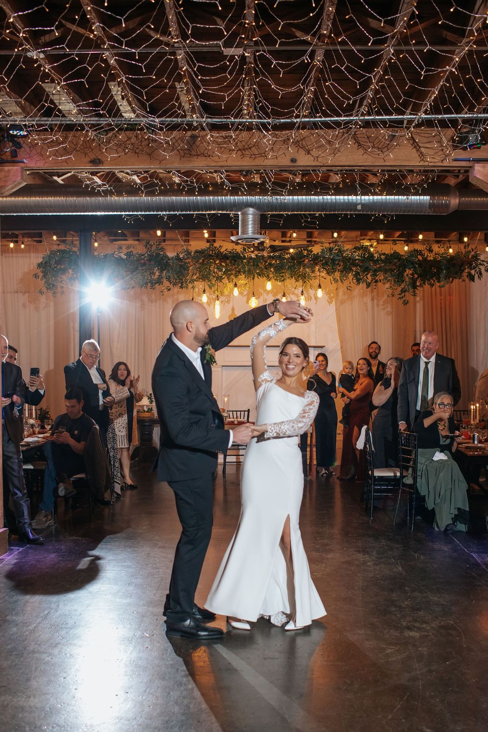 Wedding reception dance floor filled with guests under twinkling lights and greenery decor.