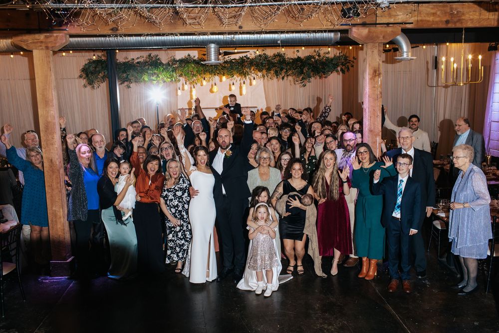 Wedding guests celebrate and cheer during reception in rustic venue with exposed beams.