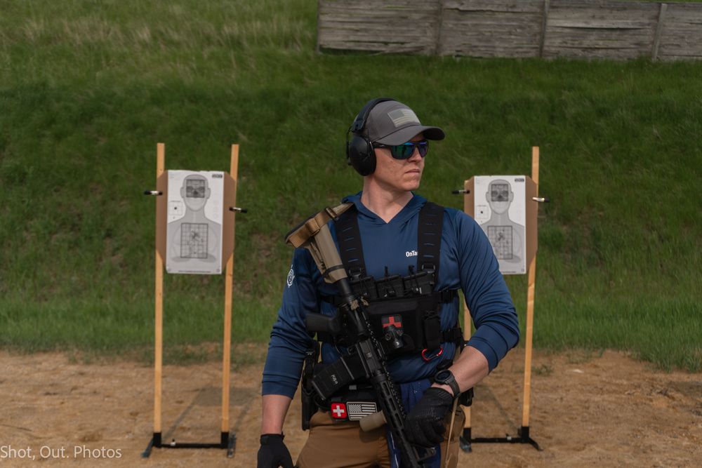 Tactical shooter in blue shirt and protective gear stands at outdoor range with target practice setup behind.
