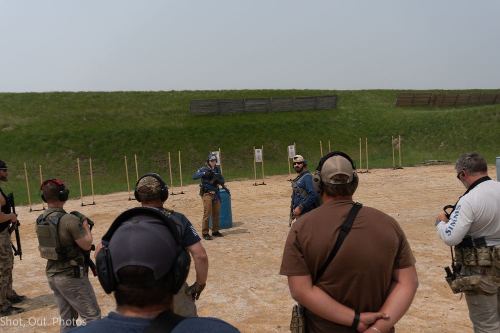 People stand in a line at an outdoor shooting range with rifles pointed downrange during target practice.