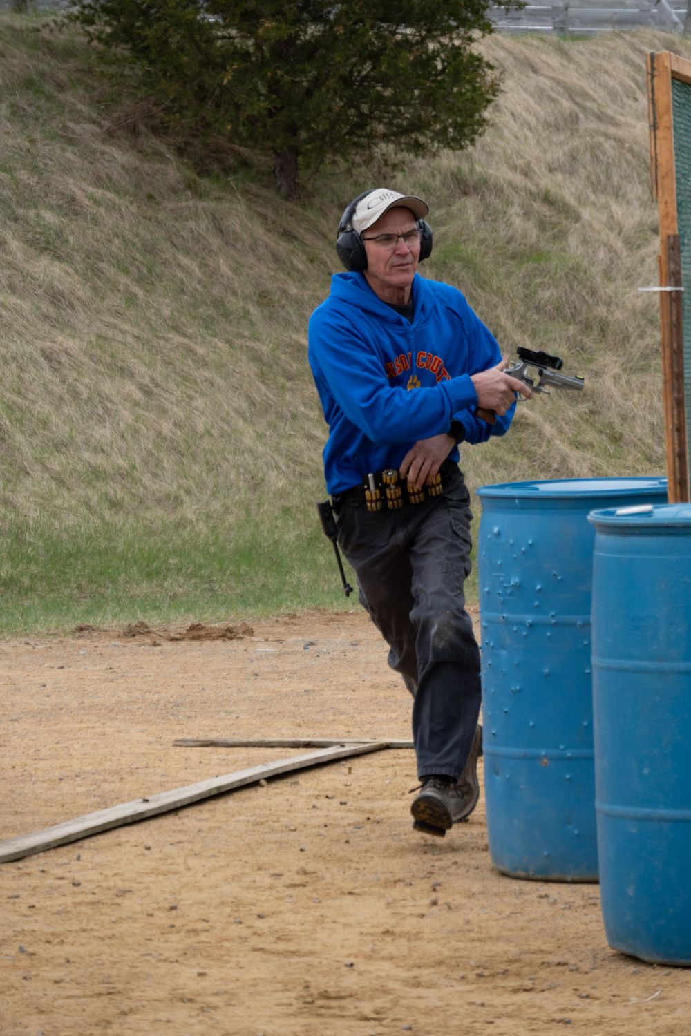 Man in blue hoodie and ear protection holds a pistol at an outdoor shooting range with blue barrels.