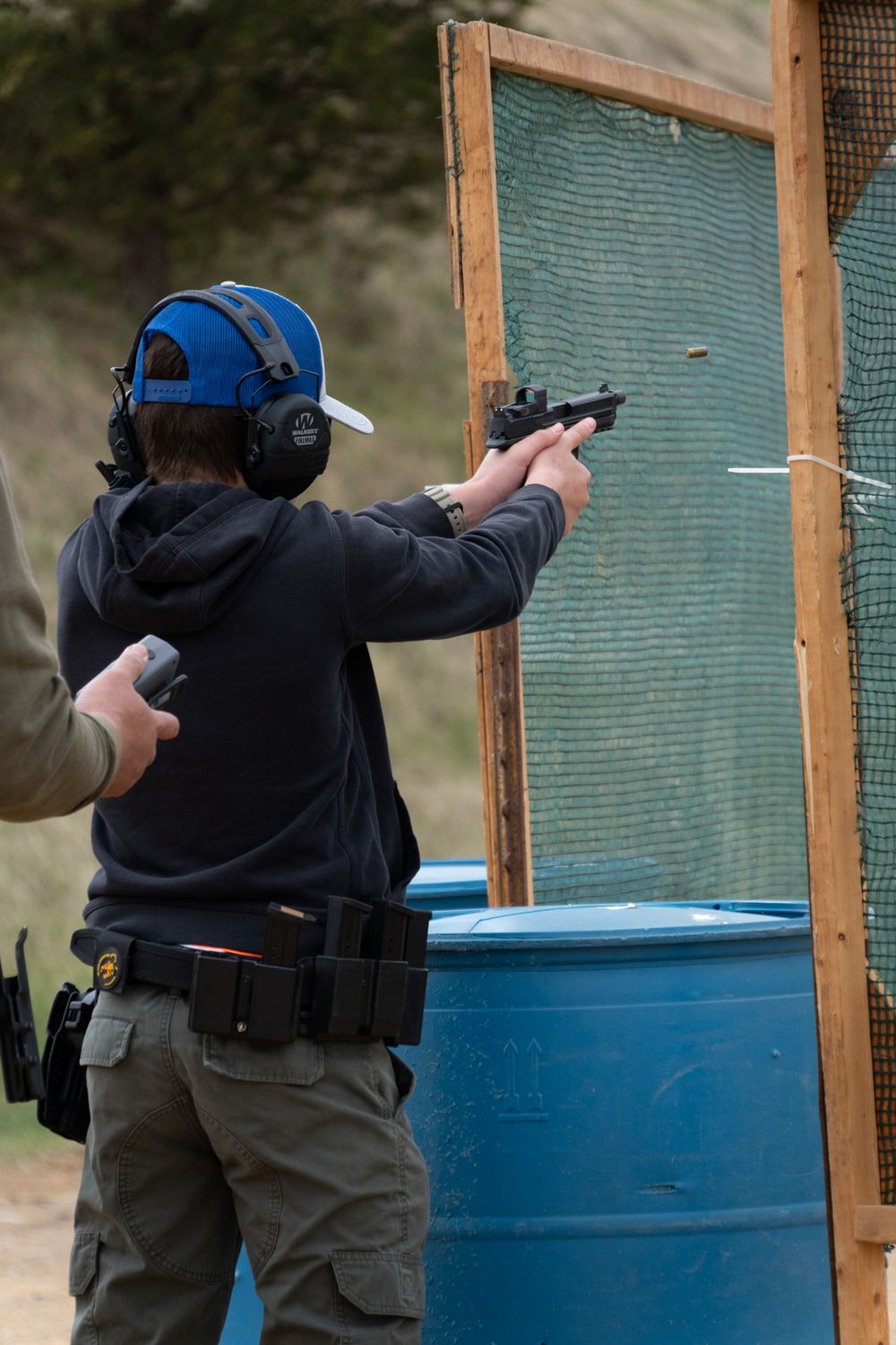 Person in blue cap and ear protection shooting a pistol at an outdoor gun range near a wooden barrier.