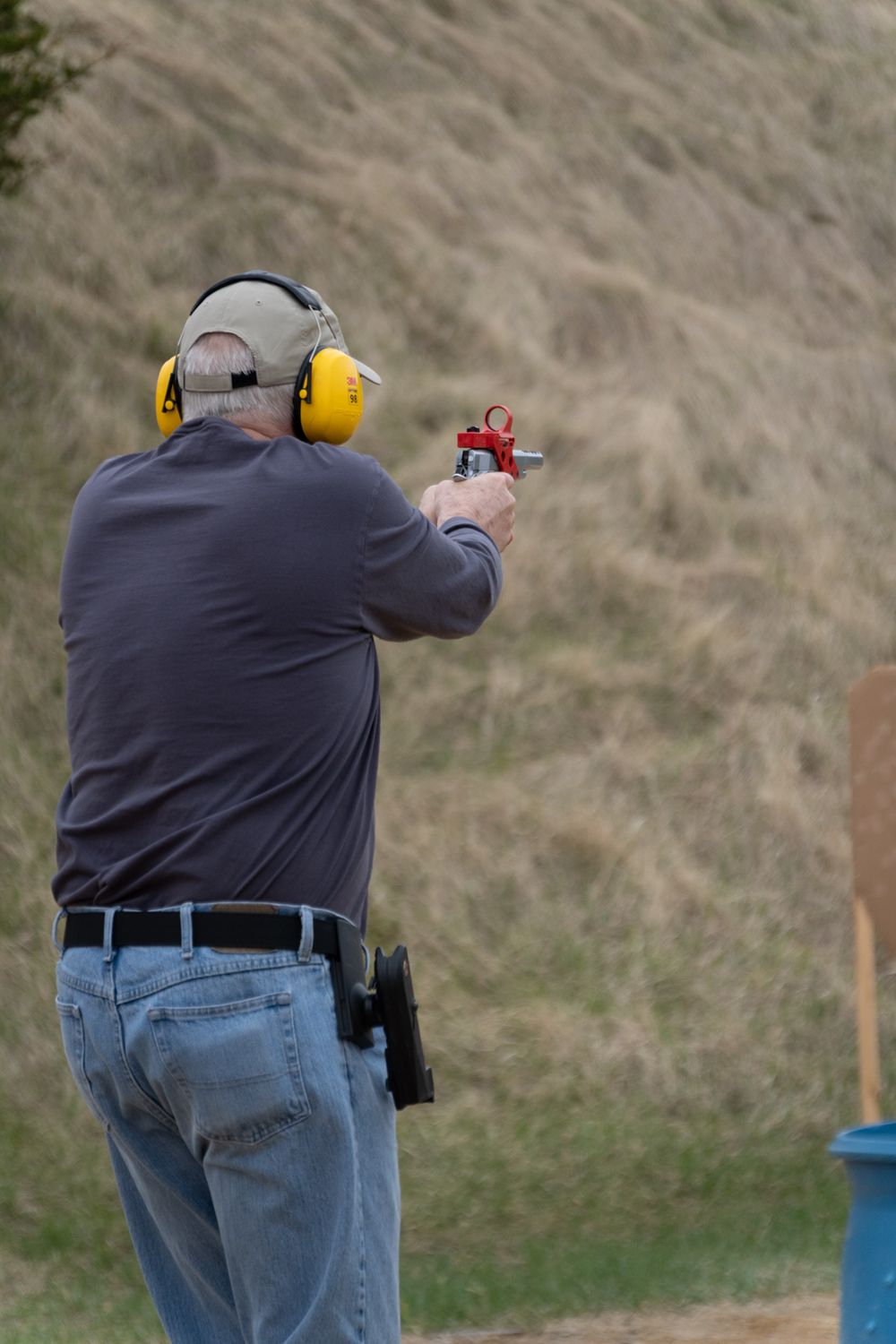 Man wearing yellow ear protection aims a red pistol at an outdoor shooting range with a grassy hillside backdrop.