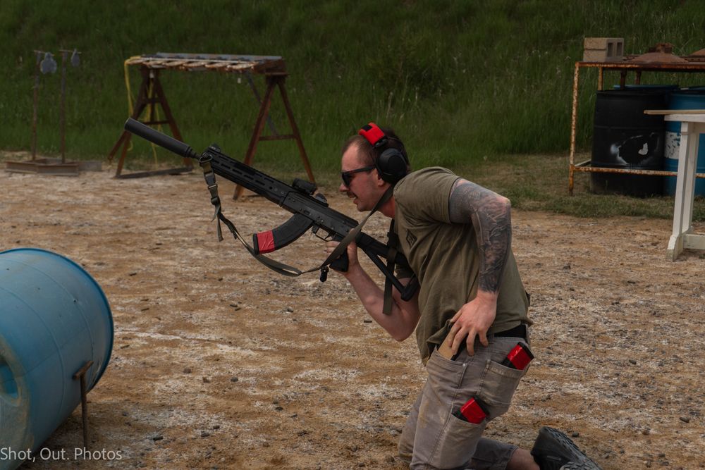 A person in red and black protective gear practices target shooting at an outdoor shooting range.