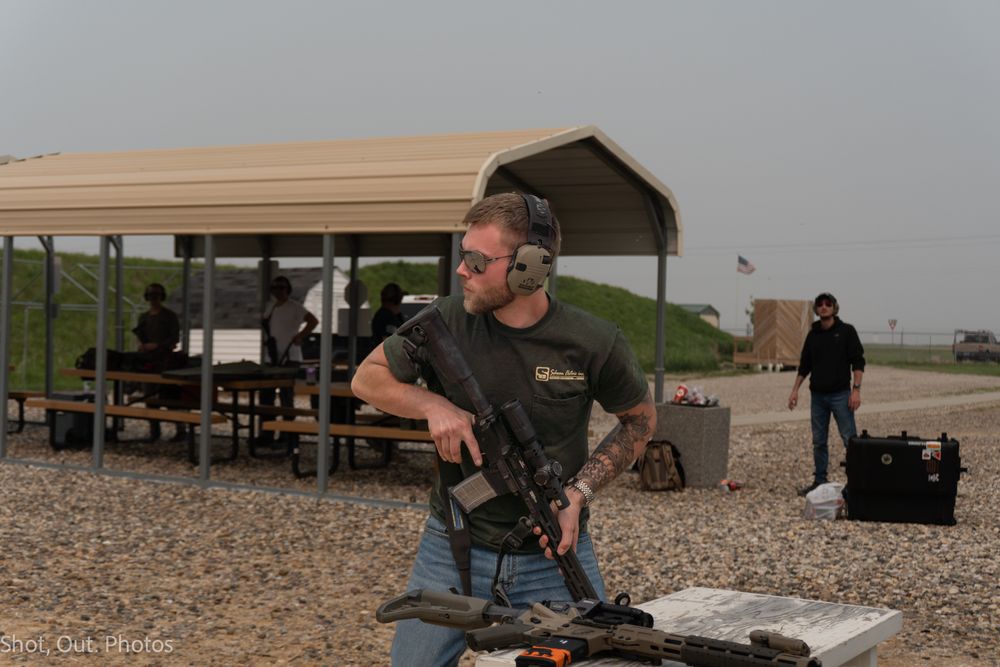 A person aims a rifle at a shooting range while others observe from a covered area during target practice.