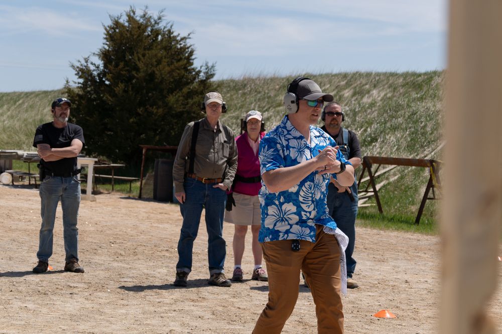 Man in floral shirt shooting at outdoor range while spectators watch on a sunny day.