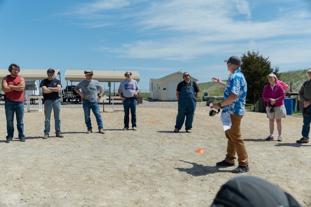 Group of people gathered outdoors at a rural location listening to a presenter during a daytime event.