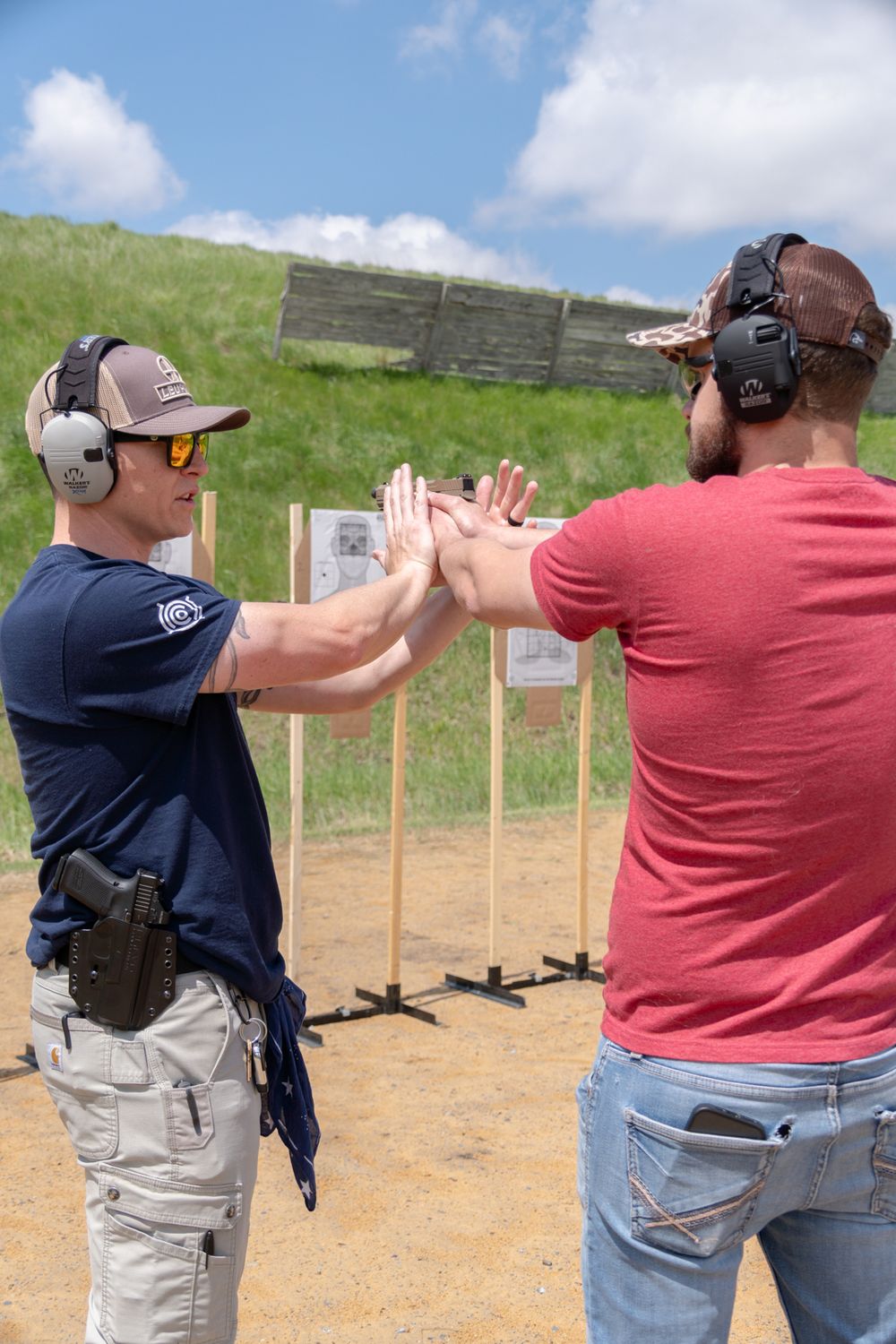 Two men in ear protection high-five at an outdoor shooting range with paper targets visible in the background.